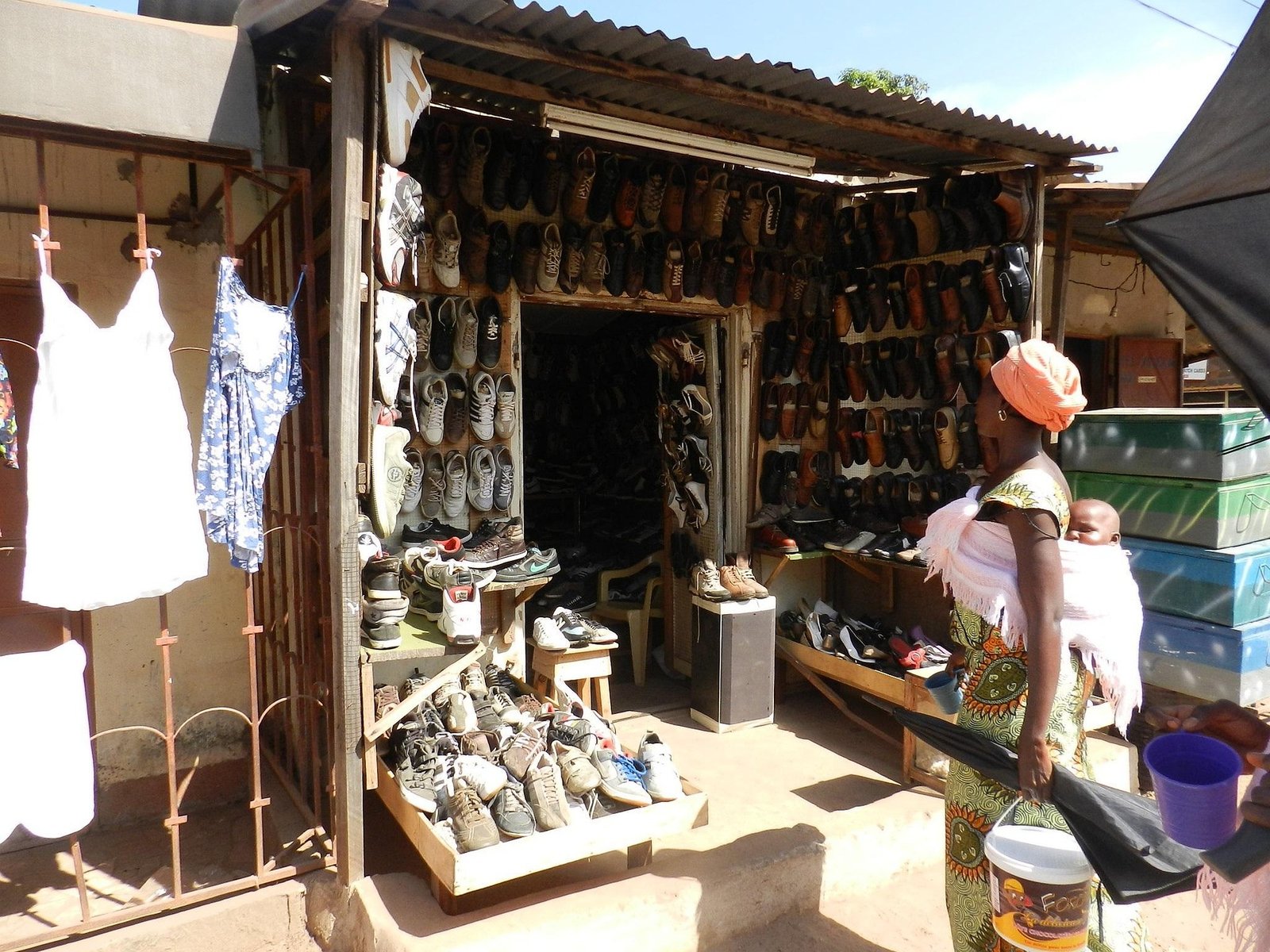 A roadside stall with rows of shoes on wooden shelves under a corrugated iron roof, with a woman in a headwrap and a baby on her back standing beside it